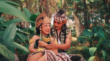 Geleneksel bir Java dans çiftinin yemyeşil bir ormanda birlikte oturduğu neşeli ve romantik bir sahne. Gülümsüyorlar, gülüyorlar, el ele tutuşuyorlar, sevgilerini ve mutluluklarını ifade ediyorlar..