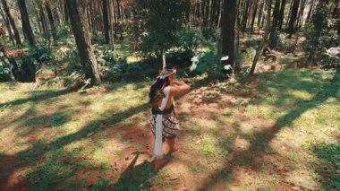 Yüksek açılı bir çekim, geleneksel bir Cava okçusunu arkadan takip eder. Poz verirken çıplak ayakla güneşli, çimenli, sakin bir çam ormanında yürür..