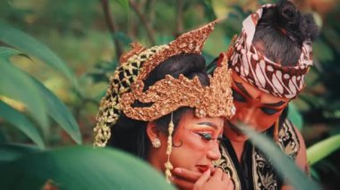 Gür yeşil yapraklarla çerçevelenmiş geleneksel bir Cava çiftinin yakın plan romantik bir sahnesi. Erkek, kadının yüzüne nazikçe dokunur şefkatli, güler yüzlü bir yakınlık ve şefkat anlarını paylaşırken..