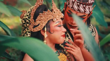 Gür yeşil yaprakların arasından görünen geleneksel bir Java çiftinin güzel, samimi bir fotoğrafı. Ormanda gizli ve romantik bir atmosfer yaratarak mutlu ve sevgi dolu bir etkileşimi paylaşıyorlar..