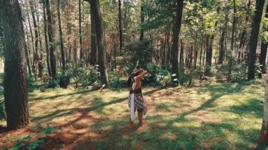 Geleneksel giyinmiş yalnız bir Javalı okçunun geniş açılı, geniş bir açılı görüntüsü. Geniş, güneşli bir orman açıklığında duruyor. Arkadan görünüyor, uzun ağaçlarla çevrili..