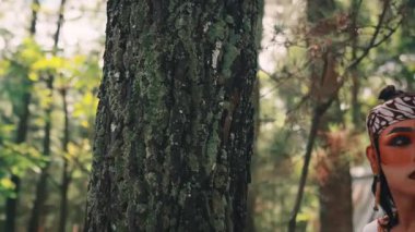 Dramatik makyajlı ve geleneksel okçu kostümlü bir Cavalı erkek dansçı ormanda bir ağacın yanında duruyor, ciddi ve tetikte bir ifadeyle etrafına bakıyor..