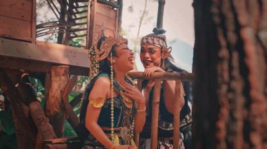Kostümlü ve makyajlı geleneksel bir Cava çifti, Endonezya folklorunun mutlu bir sahnesini anlatan ormandaki bir kır evinin balkonunda neşeli, gülen bir anı paylaşıyor..