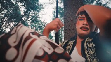 Geleneksel kostümlü bir Java dansçısı ağaca yaslanır, bitkinlik ve umutsuzluk gösterir. Güneşin orman tepe örtüsünün arkasında parladığı alçak açılı çekim dramatik ve duygusal bir sahne yaratır..