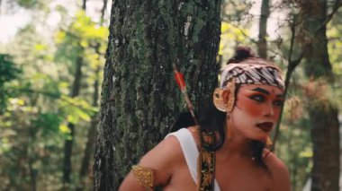 Geleneksel bir Cava okçusunu canlandıran dramatik makyajlı bir erkek dansçı, yemyeşil orman ortamında bir ağaca yaslanırken anlamlı ve kederli bir dans sergiliyor..