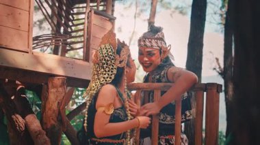 Süslü kostümlü bir erkek ve dişi Cava dansçısı ağaç evde neşeli ve samimi bir anı paylaşıyor. Gülüyorlar ve gülümsüyorlar erkeğin kadının yüzüne nazikçe dokunması gibi.