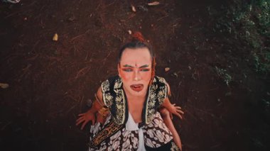 Bir Java dansçısının orman zemininde diz çöktüğü dramatik, yüksek açılı bir çekim. Umutsuz, kederli bir ifadeyle kafasını kaldırıp geleneksel tiyatro performansının güçlü bir anını yakalıyor..