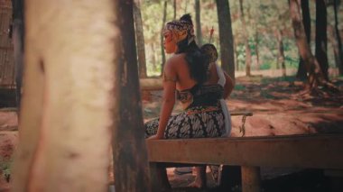 Bir ön plan ağacı tarafından çerçevelenmiş, geleneksel bir okçuyu canlandıran bir Java dansçısı, uzaklığa bakarken derin derin ve güçlü bir poz vererek ormanın bir çıkıntısında oturur..