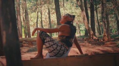 Geleneksel okçu kıyafeti giymiş bir Java dansçısı çam ormanındaki bir kirişe oturur ve gökyüzüne bakarken ihtişamlı ve düşünceli bir poz verir..