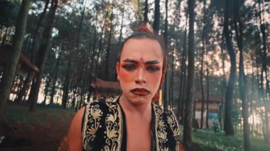 Dramatik makyajlı, ormanda oturan bir Java dansçısının düşük açılı çekimi. Kamera geri çekilirken ciddi ve yoğun bir ifadeyle doğrudan kameraya bakar ve ormanı ortaya çıkarır..