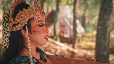 Geleneksel altın bir başlık takmış ve karmaşık makyaj yapmış bir kadın Cava dansçısının çok güzel bir profil çekimi. Düşünceli bir ifadeyle ormanın içine bakıyor..