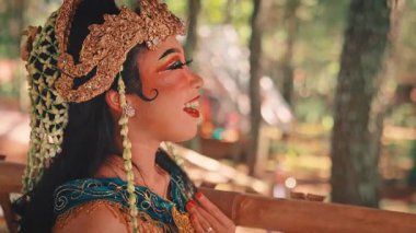 Süslü, altın başlıklı ve geleneksel makyajlı güzel bir Cava dansçısının yan görüntüsü. Orman ortamında biriyle konuşurken neşeyle gülüyor..
