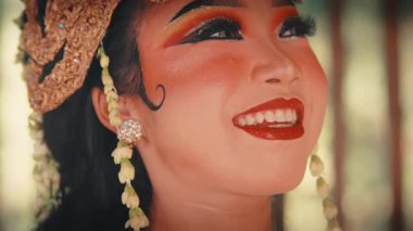 Dramatik makyaj ve altın bir başlıkla süslenmiş güzel bir Cava dansçısı yüzü. Işıl ışıl gülümser ve zarif bir şekilde yüzüne dokunur, saf mutluluğu ifade eder..