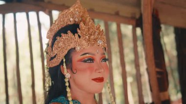Süslü altın bir başlık ve geleneksel kıyafetler giyen güzel bir Java dansçısının orta boy yakın çekimi. Zarif bir şekilde poz veriyor, etrafına sakin ve büyüleyici bir ifadeyle bakıyor..