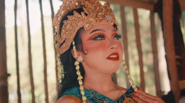 Büyüleyici altın başlık ve kostüm giymiş, zarif poz veren geleneksel bir kadın Cava dansçısı. Başlığına dokunuyor ve etrafına sakin ve güzel bir ifadeyle bakıyor..