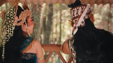 Arkadan çekilen, kostümlü geleneksel bir Java çifti ormana bakan bir balkonda oturuyor. Birbirlerine dönerler, gülümserler ve romantik bir kur sahnesinde etkileşime girerler..