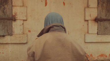 Sırtı kameraya dönük, gri kapüşonlu ve bej ceketli yıpranmış bir duvara karşı oturan, boyalı ve tahta panjurlu bir adam..