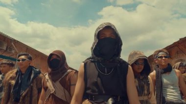 Sekiz kişilik bir grup engebeli, post-apokaliptik giyinmiş, ıssız bir şehir ortamında güvenle ayakta duruyor. Maske ve güneş gözlüğü takıyorlar, arka planda dramatik bir gökyüzü olan stil ve aksesuar karışımı sergiliyorlar..