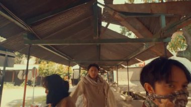 Açık hava ortamında bir barınağın altında dans eden üç kişilik bir grup. Eşsiz, dalgalı giysiler giyiyorlar, dinamik hareketler ve ifadeler sergiliyorlar. Arka planda ağaçlar ve binalar ile kırsal bir ortam yer alıyor..