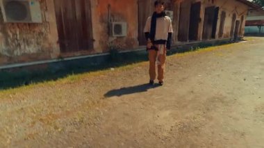 Tahta panjurları ve kliması olan eski, yıpranmış bir binanın önünde duran biri. Sahne, parlak mavi bir gökyüzünün altında nostaljik bir atmosfer yaratıyor..