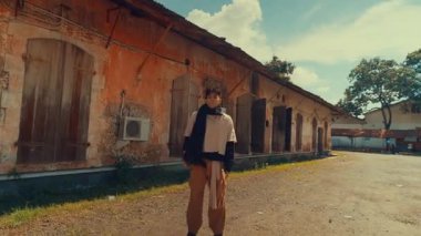 Arka planda eski binalar olan açık havada yürüyen biri. Sahnede kırsal mimari, yeşillik ve berrak mavi bir gökyüzü var, sakin bir atmosfer yaratıyor..