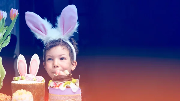 Banner with cute little boy with bunny ears looking at tasty easter sweet cake, funny kid and happy Easter holidays celebrating. Copy space. Selective focus.