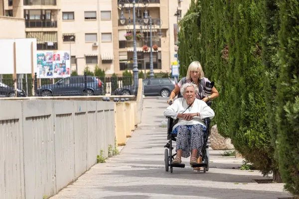 Sarışın kadın yaşlı annesini tekerlekli sandalyeyle sokağa itiyor.