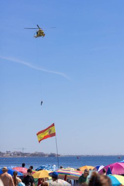 Kurtarma helikopteri havadaki dalgıcı okyanusa indirirken bir grup insan sahilden rüzgarda dalgalanan İspanyol bayrağını seyrediyor.