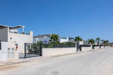 Modern beyaz evler, manzaralı bahçeler ve parlak mavi gökyüzünün altında palmiye ağaçları bulunan kapalı bir topluluk manzarası..