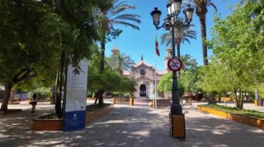 İspanyol bayrağı, sokak feneri ve Torrevieja tabelasının yer aldığı uzak bir manzaradan başlayan kamera, düzgünce Plaza de la Constitucin 'e doğru ilerliyor.