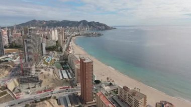 Benidorm sahil şeridinin yüksek açılı görüntüsü kumlu plajlar, modern gökdelenler ve bulutlu bir gökyüzünün altındaki dağları çevreleyen