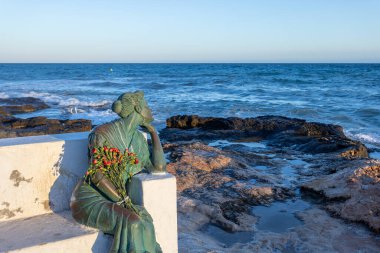 Torrevieja 'daki Bella Lola heykeli, elinde kırmızı çiçekler, denize bakıyor, geri dönmeyen denizciler için beklemeyi ve kaybetmeyi simgeliyor.
