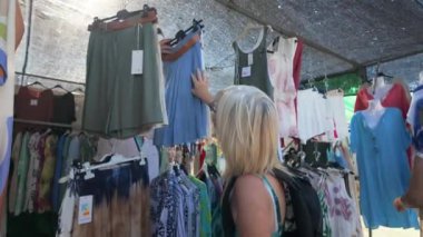 Bir kadın, Torrevieja 'nın açık hava pazarındaki bir giyim mağazasında etek ve şortlarını inceliyor, gölgeli bir gölgelik altında çeşitli moda eşyalarına göz atıyor.