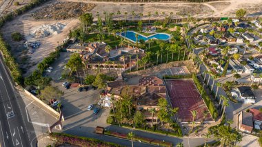 İspanyol kırsalındaki bir tatil köyünün insansız hava aracı görüntüsü. Yüzme havuzu, tenis kortu ve yemyeşil bir çevresi var..