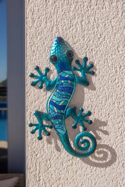 Vibrant blue mosaic gecko wall decoration casting a shadow on a textured beige wall, illuminated by bright sunlight. clipart