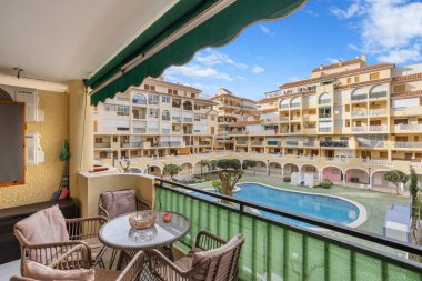 Cozy balcony with wicker furniture overlooking a Mediterranean-style residential complex featuring a communal swimming pool and vibrant architecture. clipart