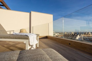 Relaxing rooftop terrace with a sun lounger, straw hat, and glass railing offering expansive views of the surrounding area. clipart