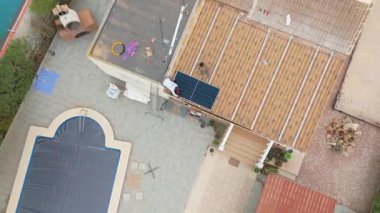 Bahçeler ve yüzme havuzlarıyla çevrili, döşemeli bir çatıya güneş panelleri kuran işçilerin olduğu bir konutun tepeden aşağı insansız hava aracı görüntüsü..