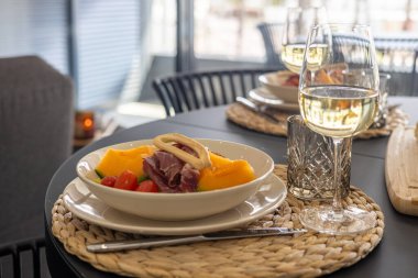 Bir tabak kavun, prosciutto, kiraz domatesleri ve modern, rahat bir ortamda beyaz şarap içeren bir yemek masasının yakın çekimi..