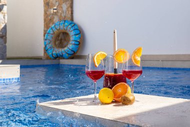 Bardaklar dolusu sangria ve taze narenciye meyveleri bir havuzda, su sıçramasıyla çevrili bir platforma yerleştirilmiş. Canlandırıcı bir sahne. Yaz yaşam tarzı görüntüsü için mükemmel..