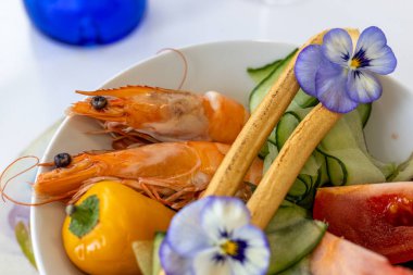 Renkli karidesli karides salatası, sarı dolmalık biber, salatalık kurdelesi, ekmek çubukları ve canlı yenilebilir çiçekler. Görsel olarak çekici bir deniz ürünleri tabağı beyaz bir kasede servis edilir, yemek sunumları için mükemmeldir..