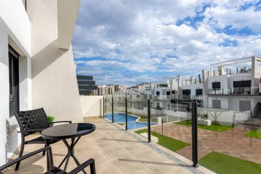 Geniş bir teras, siyah sandalyeli ve masa manzaralı modern bir apartman kompleksi ve parçalı bulutlu bir gökyüzünün altında yüzme havuzu..