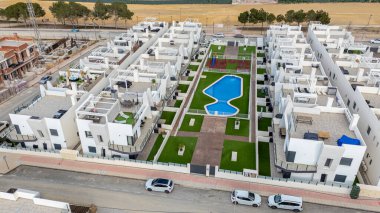 Beyaz binalar, yeşil alanlar ve merkez havuz içeren modern bir konut kompleksinin insansız hava aracı görüntüsü..