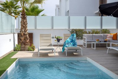 Özel havuz kenarında iki bej şezlonglu, tropik havlusu, şarap bardakları ve bir palmiye ağacı var..
