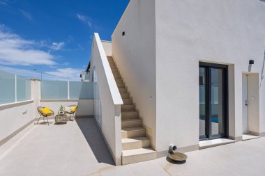 Çatıya merdiven erişimi olan geniş fayanslı teras. Sarı yastıklı ve açık hava dekorlu modern oturma alanı..