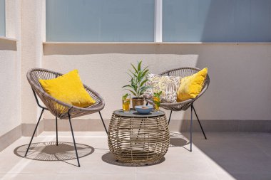 Sarı yastıklı iki yuvarlak sandalye, bir hasır masa, içecekler ve bir bitki süsü içeren şık ve rahat bir teras düzeneği..