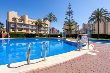 Güneşli bir günde palmiye ağaçları ve modern dairelerle çevrili paslanmaz çelik basamakları ve havuz kaldırma havuzu olan yüzme havuzunu davet etmek..