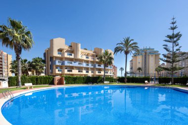 Uçsuz bucaksız yuvarlak havuz mavi gökyüzünü yansıtıyor, arka planda güneşli apartmanlar ve tropikal ağaçlar var..