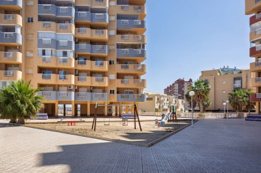 Balkonlu uzun turuncu bir apartman ve çocuklar için kumlu bir oyun alanı..