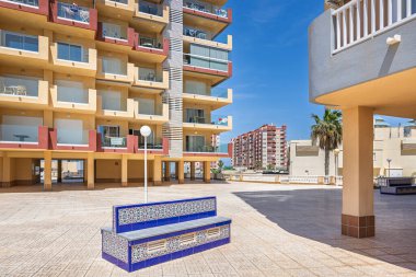 Modern cam önlü balkonlar ve sıcak tonlu binalarla çevrili fayanslı bir avluda renkli seramik bank..